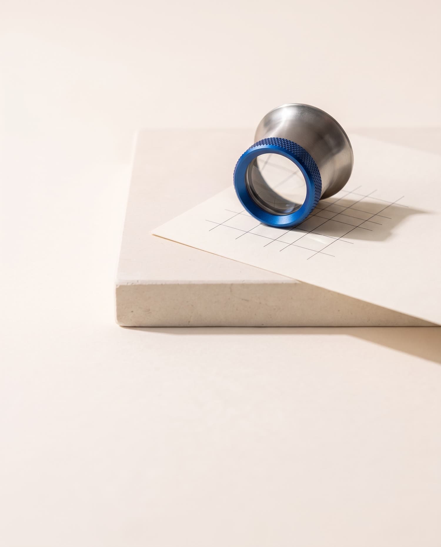 A steel jeweller's loupe with a cobalt ring resting on a sheet of grid paper, on a stone plinth